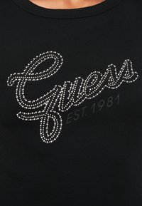 Czarny prążkowany top na ramiączkach z logo "Guess" wykonanym z kryształków, z nadrukiem "EST. 1981" poniżej w subtelnym ciemnym kolorze.