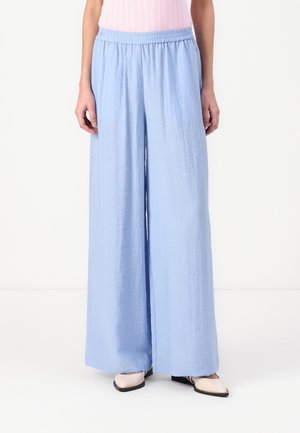 Vrouw draagt lichtblauwe wijde broek met elastische taille en een lichtroze mouwloos topje, gecombineerd met beige puntige schoenen.
