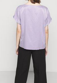 Persona di spalle che indossa una blusa a maniche corte lavanda e pantaloni neri larghi su uno sfondo chiaro e semplice.
