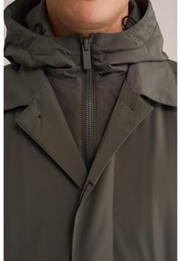 Pilka zip-up striukė su aukštu apykakle ir kepure. Audinys lygus, su matomomis siūlėmis ir ryškiu priekinio užtrauktuko uždarymu.