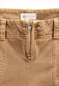 Pantalon en denim beige avec une fermeture éclair, des coutures apparentes et une étiquette indiquant "GAP SANDY LIANG" en haut. Tissu texturé.