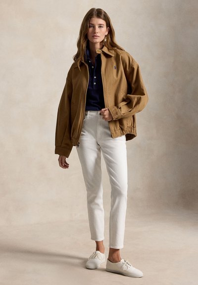 Vrouw draagt een bruin jasje met rits, marineblauw poloshirt, witte broek en witte sneakers, staand tegen een effen beige achtergrond.