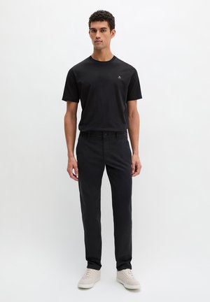 Mann steht und trägt ein schwarzes kurzärmeliges T-Shirt, schwarze Hose und hellbeige Sneakers vor neutralem weißen Hintergrund.