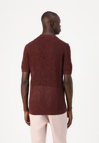 Polo en dentelle bourgogne à manches courtes, avec un design texturé et un col et des poignets côtelés. Détail subtil en maillage ouvert.