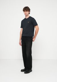 Črna bombažna majica s kratkimi rokavi in okroglim izrezom, z malim rumenim logotipom. Kombinirana s temno modrimi kavbojkami in črnimi čevlji.