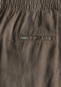 Brun tyg med en skrynklig struktur, med en elastisk midjeband och en liten metallisk etikett som är sydd på bakfickan.