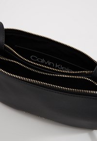 Czarna torebka ze skóry naturalnej z elementami zamka błyskawicznego w kolorze złotym, dwoma przegródkami oraz gładkim wnętrzem. Oznaczona napisem "Calvin Klein" wewnątrz.