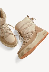 Bottes isolées beige avec doublure en fourrure douce, accents en suède texturé, lacets élastiques et semelle en caoutchouc présentant un motif surélevé et un profil orangé.