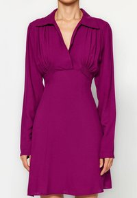 Lila klänning med krage, långa ärmar och rynkdetaljer vid midjan. Tillverkad av ett mjukt tyg, har den en utsvängd silhuett.