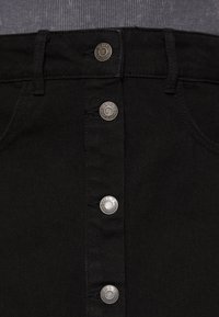 Lähikuva mustast denim-seeliku etupinnast, jossa on neliä hopeanväristä merkkilogolla varustettua nappia ja vyölenkkejä, yhdistettynä harmaaseen ribbipaitaan.