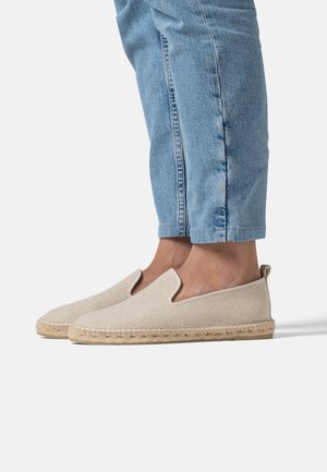 Personne portant des espadrilles beige clair sans lacets avec semelles tissées et un jean bleu clair retroussé, debout sur une surface blanche.