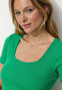 Haut vert à col rond, à manches courtes et côtelé. Le tissu est lisse avec une silhouette ajustée. Le modèle porte des boucles d'oreilles créoles.
