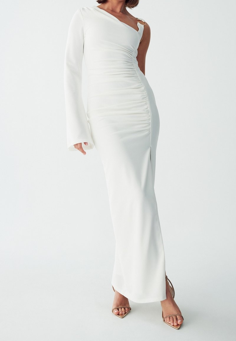 Femme portant une longue robe blanche avec une manche longue et une épaule dénudée, associée à des talons ouverts dorés.