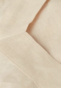 Béžová lněná tkanina s texturovanou vazbou a viditelnými vzory vláken, s pečlivě prošívanými okraji, vytvářející přirozený vzhled.