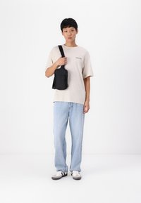 Crna crossbody torba s jednim remenom, nošena preko svijetlosmeđe košulje. Plave široke traperice i sive tenisice s crnim detaljima.