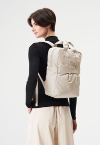 Helles beiges Rucksack aus glattem Material, ausgestattet mit zwei Oberhandgriffen, gepolsterten Schulterträgern und einer Fronttasche mit minimalistischen Akzenten.