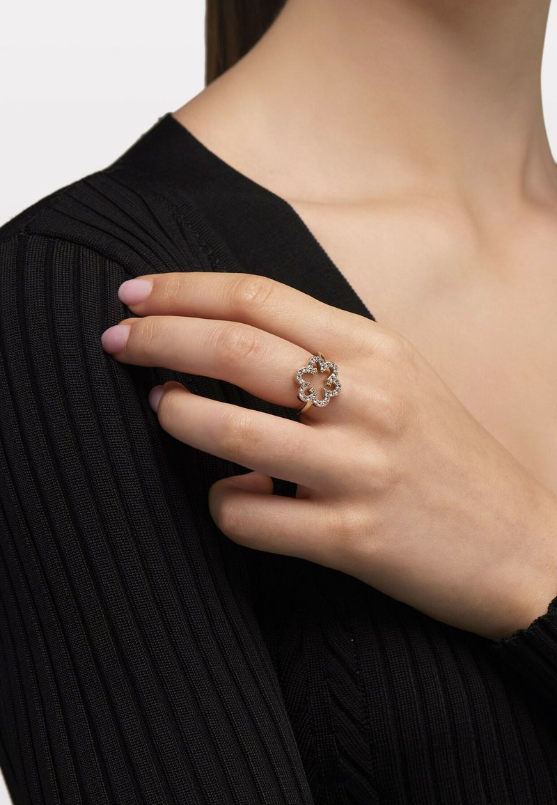Anello argento a forma di fiore con pietre trasparenti, montato su una fascia liscia. La mano poggia su un tessuto a costine nero.