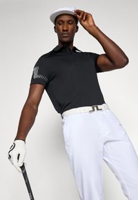 Camisa polo preta com um padrão xadrez na manga, combinada com calças brancas e um boné branco. Segurando um taco de golfe enquanto ajusta o boné.