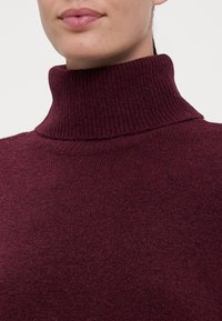 Burgundy kötött garbó pulóver bordás textúrával, amely kiemeli a testhez simuló fazont és a magas gallért. Puha anyag finom varrási részletekkel.
