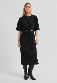 Robe midi noire en tissu doux avec un détail twist à la taille, accentuée par un bouton en métal doré circulaire et des manches courtes.
