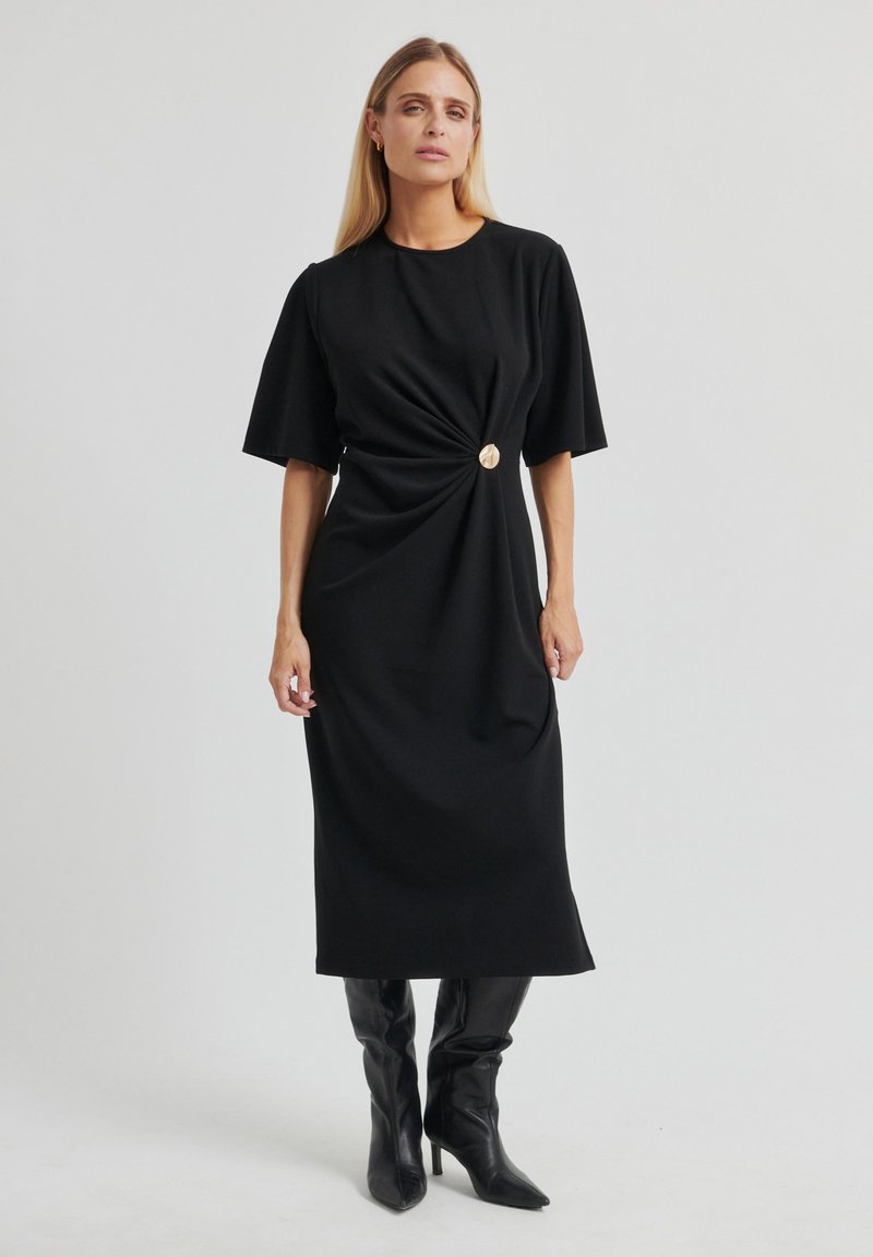 Robe midi noire en tissu doux avec un détail twist à la taille, accentuée par un bouton en métal doré circulaire et des manches courtes.