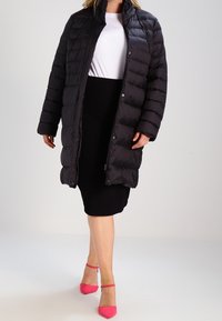 Nainen, joka pukeutunut mustaan tikattuun pitkään takkiin valkoisen topin ja mustan hameen päällä, kävelee kirkkaan vaaleanpunaisissa nilkkahihkaisissa korkokengissä yksiväristä taustaa vasten.