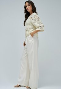 Häkeloberteil in Off-White mit weitem Ärmel und einem komplizierten Blumenmuster, kombiniert mit hochtaillierten, weißen, weiten Hosen.