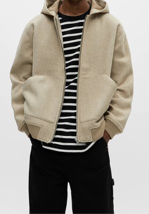 Person iført en beige hættejakke med lynlås over en sort og hvid stribet skjorte med sorte bukser, hænder delvist i jakkelommer.