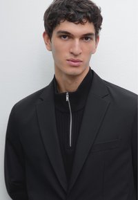 Blazer nero sopra un maglione nero a coste con zip. Il blazer presenta una tasca singola e una vestibilità su misura, con un colletto strutturato.