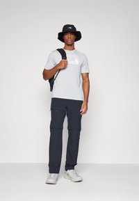 T-shirt gris clair avec un logo texturé, chapeau de seau noir, pantalon cargo noir avec zippers et chaussures de sport blanches.