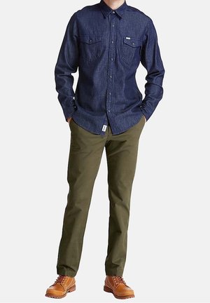 Homme portant une chemise bleu foncé à boutons, un pantalon vert olive et des bottes en cuir marron à lacets, debout les mains dans les poches.