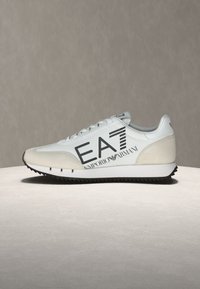 Miesten lenkkikenkä, jossa vaaleanharmaa päällinen, mokkasisustukset, mustat logokirjaimet, litteät nauhat ja teksturoitu musta kumipohja.