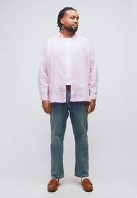 Άνδρας με πλεξούδες, που φοράει ροζ ανοιχτό ριγέ πουκάμισο, λευκό μπλουζάκι, μπλε τζιν και καφέ μοκασίνια, στέκεται μπροστά σε απλό ανοιχτό φόντο.