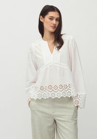 Blusa bianca a maniche lunghe con scollatura a V, caratterizzata da pieghe verticali, vita arricciata e bordo in pizzo floreale.