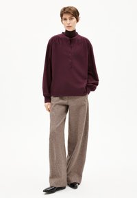 Blusa bordeaux a maniche lunghe con colletto alto e dettagli con bottoni, abbinata a pantaloni grigi larghi e strutturati e scarpe nere.