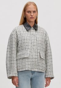 Tweed jasje in wit en zwart met een zwarte leren kraag, met een boxy vorm en twee voorkruiszakken. Gelaagd stof met een ruitpatroon.