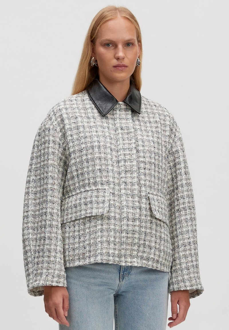 Tweed jasje in wit en zwart met een zwarte leren kraag, met een boxy vorm en twee voorkruiszakken. Gelaagd stof met een ruitpatroon.