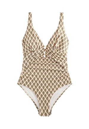 Kvinde med solbriller og hestehale iført en mønstret ét-stykke badedragt, der står ved roligt hav under klar himmel.