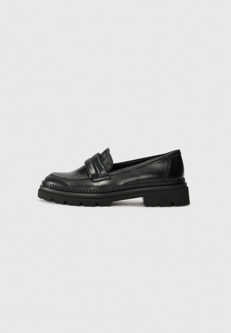 Mocassin en cuir noir avec semelle en caoutchouc épaisse et détail de surpiqûres autour de la partie supérieure, présenté de profil sur un fond gris uni.