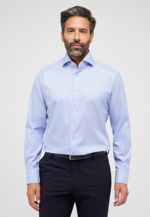 Light blue long-sleeve shirt with a small dotted pattern, spread collar, and button closure. Worn with dark trousers, featuring a sleek belt.