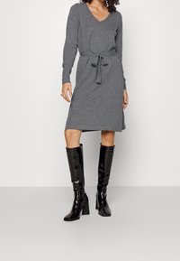 Abito grigio a maniche lunghe con scollo a V e vita legata. Abbinato a stivali neri in pelle laccata al ginocchio con tacco a blocco.