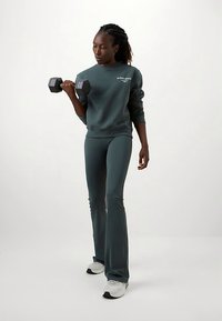 Άτομο με σκούρο πράσινο φούτερ και παντελόνι σηκώνει ένα μαύρο αλτήρα με το δεξί χέρι, στέκοντας σε λευκό φόντο.