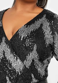 Nära bild av en kvinna som bär en svart och silver paljett-topp med djup V-ringning, som visar upp dess glittrande mönster och textur.