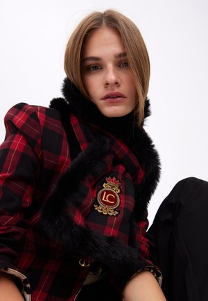 Chaqueta de cuadros en rojo y negro, con un cuello de piel sintética negra y un emblema LC ornamentado con detalle de corona. Textura de tela suave.