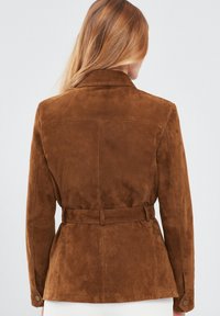 Bruine suède jas met een ceintuur in de taille, getailleerde pasvorm en een puntige kraag. Kenmerkt zich door een gladde textuur en heeft geen zichtbare patronen of versieringen.