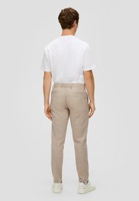Man staat met zijn rug naar de kijker, gekleed in een wit T-shirt, beige broek en witte sneakers tegen een effen lichte achtergrond.