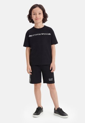 Dieťa s vlasmi po ramená nosiace čierne tričko EA7 Emporio Armani, zodpovedajúce šortky a čierne tenisky, stojace na bielom pozadí.