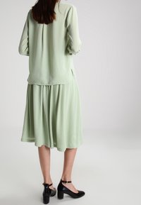 Hellgrünes, plissiertes Midikleid mit fließendem Schnitt, kombiniert mit schwarzen Ankle-Strap-Absätzen. Glatte Textur und entspannter Sitz.