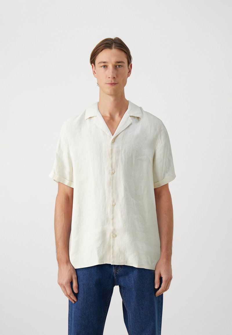Hellbeige Kurzarm-Leinenshirt mit ausgeschnittenem Kragen und fünf Knöpfen, kombiniert mit blauen Jeans. Feine Textur sichtbar.