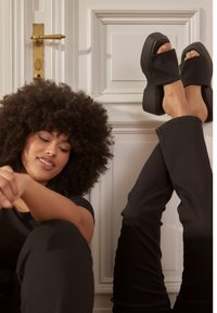 Sandálias de plataforma pretas com uma tira larga e textura suave, mostradas em pés numa posição relaxada. Calças escuras são usadas, criando contraste.